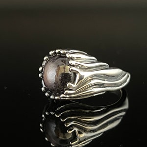 Może przedstawiać: Srebrny pierścionek z dużym, okrągłym, ciemnofioletowym kamieniem szlachetnym osadzonym w teksturowanym wzorze w kształcie dłoni.