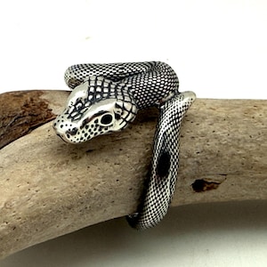May include: A silver-toned ring shaped like a coiled snake with a textured, scale-like pattern across its body. The snake head features dark, recessed detailing around the eyes and snout, creating a high-contrast look against the polished metal.