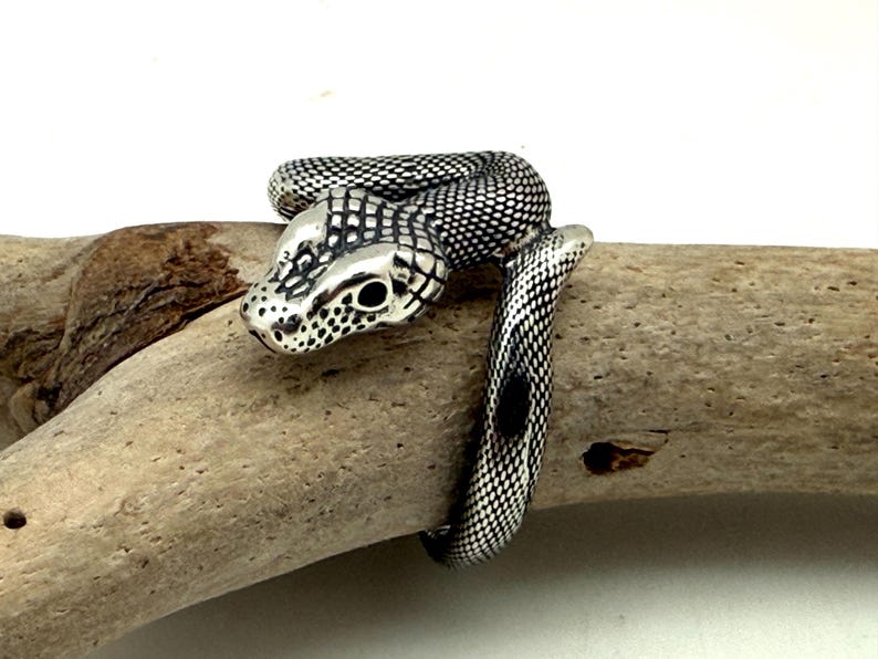 May include: A silver-toned ring shaped like a coiled snake with a textured, scale-like pattern across its body. The snake head features dark, recessed detailing around the eyes and snout, creating a high-contrast look against the polished metal.