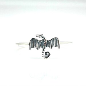 May include: A silver-colored ring featuring a detailed dragon design. The dragon has outstretched wings and a curled tail. The ring is set against a white background, highlighting the intricate details of the fantasy-themed jewelry.