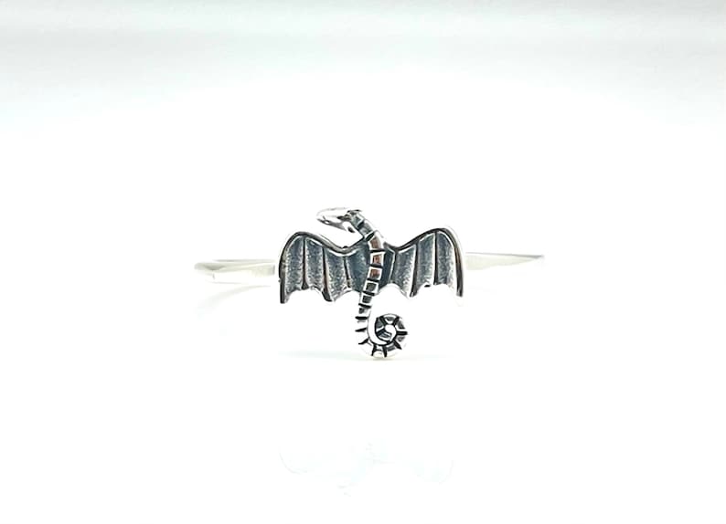 May include: A silver-colored ring featuring a detailed dragon design. The dragon has outstretched wings and a curled tail. The ring is set against a white background, highlighting the intricate details of the fantasy-themed jewelry.