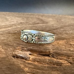 Könnte beinhalten: Ein silberner Ring mit detailliertem floralem Design. Der Ring hat ein zentrales Sonnenmuster, umgeben von kleineren floralen und perlenbesetzten Akzenten. Der Ring ist etwa 0,6 cm breit.