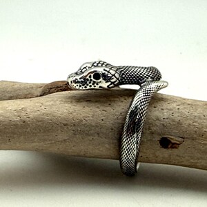 May include: A silver-toned metal ring shaped like a coiled snake. The ring features a detailed, textured scale pattern across its surface, with dark, recessed areas highlighting the snake's head, eyes, and body segments as it wraps around.