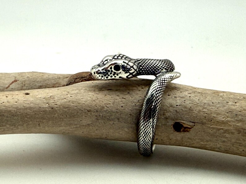 May include: A silver-toned metal ring shaped like a coiled snake. The ring features a detailed, textured scale pattern across its surface, with dark, recessed areas highlighting the snake's head, eyes, and body segments as it wraps around.