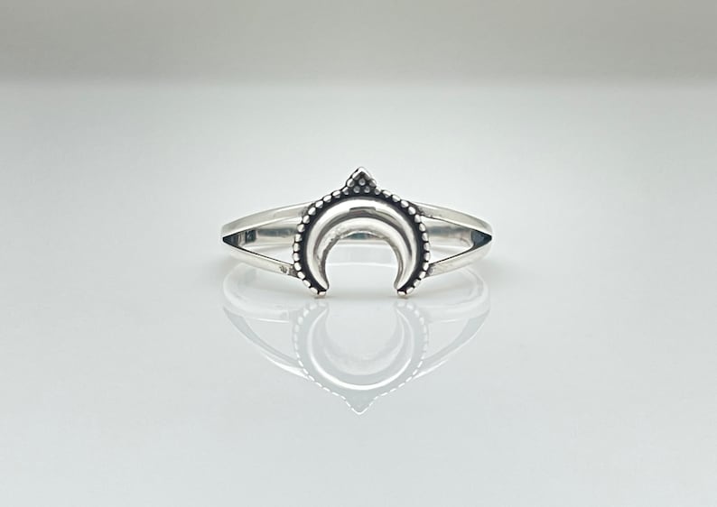 Peut inclure: Bague argent&eacute;e orn&eacute;e d'un croissant de lune. La bague pr&eacute;sente un croissant de lune d&eacute;taill&eacute; avec une bordure perl&eacute;e et un petit triangle au sommet. L'anneau est divis&eacute;, cr&eacute;ant un look unique et &eacute;l&eacute;gant.