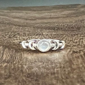 May include: A silver ring featuring a central, round, light-colored stone, flanked by crescent moon designs. The ring is set against a textured, wood-grain background, highlighting the jewelry's celestial theme. The ring's design is delicate and detailed.