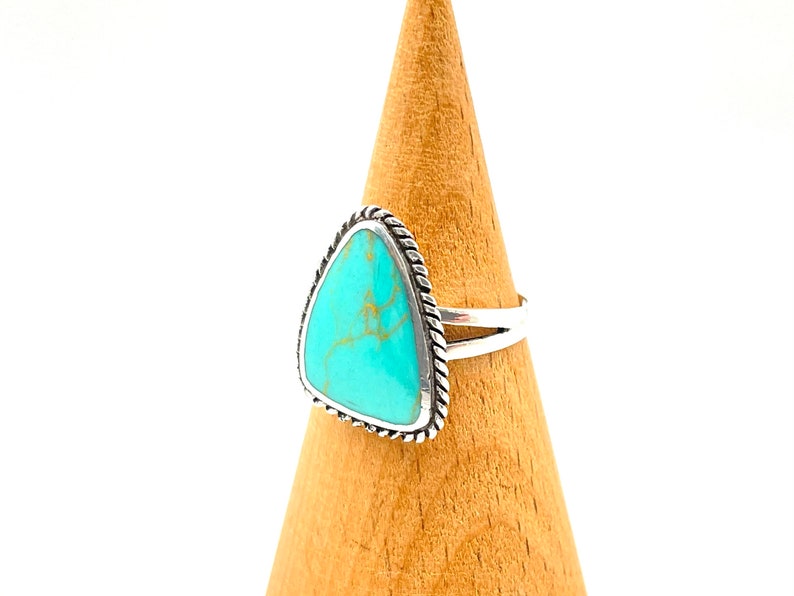 May include: A silver ring with a turquoise stone set in a triangular shape. The ring has a rope-like design around the edge of the stone.