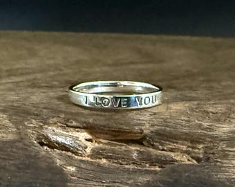 May include: A silver ring with the words "I LOVE YOU" engraved around the band. The ring is set against a rustic wooden surface, creating a contrast between the polished metal and the natural texture of the wood.