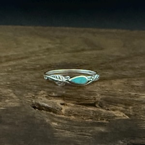 Puede incluir: Un anillo de plata con una piedra turquesa en forma de lágrima. El anillo presenta un diseño de hoja en un lado y pequeños elementos decorativos alrededor de la piedra. El anillo está sobre un fondo de madera oscura.