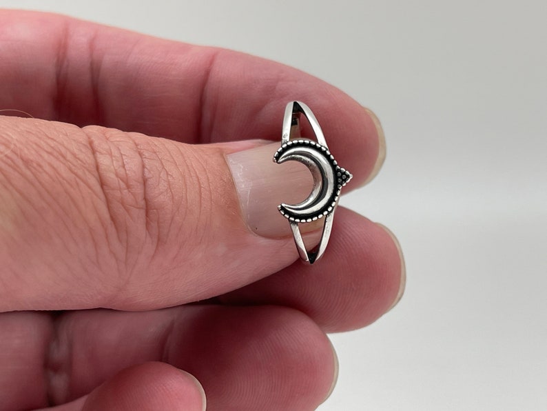Peut inclure: Une bague argent&eacute;e orn&eacute;e d'un croissant de lune. La lune est orn&eacute;e d'une bordure pointill&eacute;e et se trouve entre deux bandes parall&egrave;les. La bague a un th&egrave;me minimaliste et c&eacute;leste.