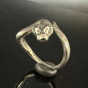 May include: A metallic ring shaped like a coiled snake with a textured, scale-like surface. The snake head is positioned at the top, facing forward with visible eyes and snout details, while the body forms the circular band of the ring.