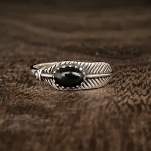 Op de afbeelding: Een zilveren ring met een zwarte ovale edelsteen gezet in een veermotief. De ring heeft een gedetailleerd, getextureerd uiterlijk, met de aderen van de veer duidelijk zichtbaar. De ring wordt gepresenteerd op een donker, getextureerd oppervlak.