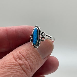 May include: A silver ring featuring an oval, blue opal stone. The opal is set in a decorative, filigree-style setting. The ring's band is a simple, polished silver. The ring is held up against a plain background.