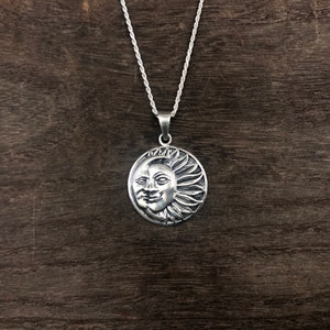 May include: A silver necklace with a round pendant featuring a detailed sun and moon design. The pendant has a face with a smile on the sun side and a crescent moon on the other side.