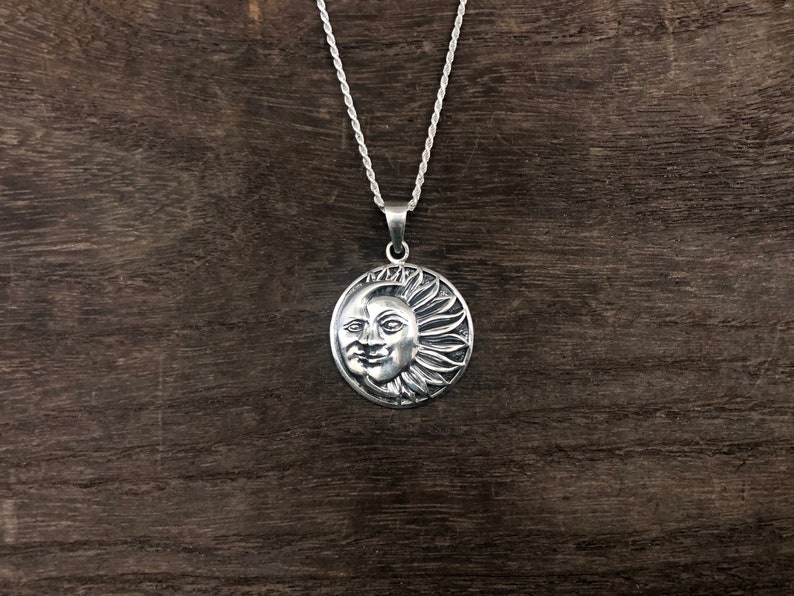 May include: A silver necklace with a round pendant featuring a detailed sun and moon design. The pendant has a face with a smile on the sun side and a crescent moon on the other side.