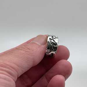 May include: A silver ring with an intricate, swirling design. The ring is wide and has a polished finish.