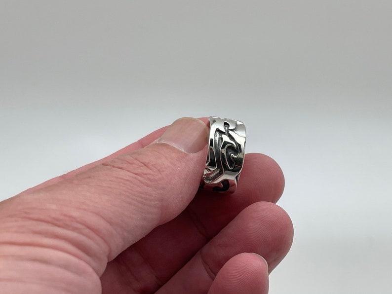 May include: A silver ring with an intricate, swirling design. The ring is wide and has a polished finish.