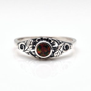 Op de afbeelding: Een zilveren ring met een ovale, donkere edelsteen in het midden. De ring heeft blad- en krulwerkontwerpen op de band. De edelsteen heeft een mix van donkerrode, bruine en groene kleuren. De ring staat tegen een witte achtergrond.