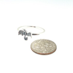 May include: A silver dragon ring with detailed wings and a curled tail. The ring is next to a dime for size comparison. The dragon is facing left, with its wings spread. The ring is on a white background.