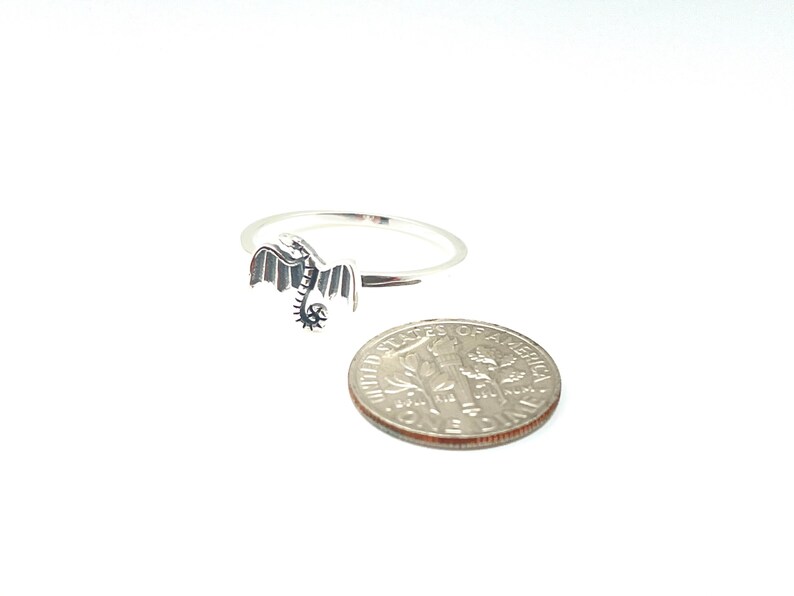 May include: A silver dragon ring with detailed wings and a curled tail. The ring is next to a dime for size comparison. The dragon is facing left, with its wings spread. The ring is on a white background.