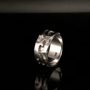 May include: A silver ring with a textured, wave-like design. The ring is wide and has a polished finish.