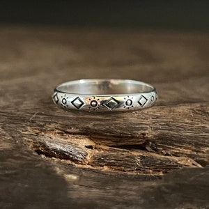 May include: A silver ring with a repeating pattern of sun and diamond shapes. The ring is set against a dark wood grain background, highlighting the ring's design. The ring is a simple band style.