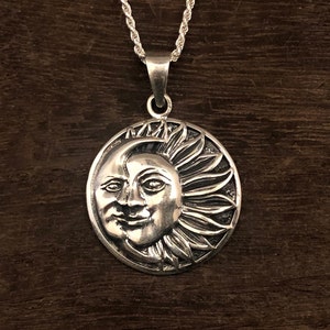 May include: A silver pendant necklace featuring a detailed sun and moon design. The sun has a face and rays, while the moon is a crescent shape.