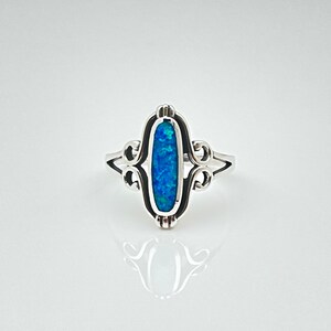 May include: A silver ring with an oval, blue opal stone. The opal is set in a decorative frame with scrollwork on the sides. The ring is on a white background.