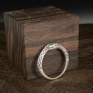 Könnte beinhalten: Ein silberner Ring mit Schlangenmuster. Der Ring hat einen detaillierten Schlangenkopf und einen strukturierten Körper, wobei der Schlangenkopf die Oberseite des Rings bildet. Der Ring wird auf einem dunklen Holzblock präsentiert.