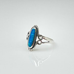 May include: A sterling silver ring featuring an oval, blue opal stone. The opal is set in a decorative bezel with scrollwork on the sides. The ring has a split shank design and is displayed on a white surface.
