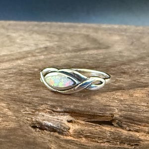 Puede incluir: Un anillo de plata con una piedra central ovalada de ópalo iridiscente. La banda presenta un diseño entrelazado. El anillo está sobre un fondo de madera natural, resaltando los detalles de la joya.