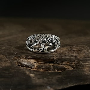 May include: A silver ring with a detailed wolf design. The wolf is depicted in a running pose, with its head turned to the side. The ring is oval-shaped and has a smooth, polished finish.