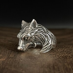 May include: A silver wolf head ring with a detailed design. The ring is made of sterling silver and features a realistic wolf head with sharp teeth and a fierce expression.