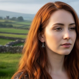 May include: Silver Celtic knot earrings. The earrings are a small, dangling design with a traditional Celtic knot symbol. The image shows the earrings worn by a person with long, red hair, set against a blurred green landscape.