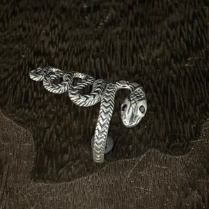 Könnte beinhalten: Ein silberner Schlangenring mit detaillierten Schuppen und geschwungener Form. Der Schlangenkopf befindet sich oben, mit Augenhöhlen. Der Ring ist vor einem dunklen, strukturierten Hintergrund platziert.