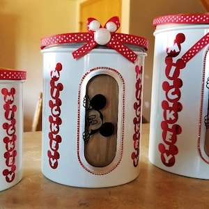 May include: Three white canisters with red polka dot lids and Mickey Mouse designs. The canisters have clear windows with Mickey Mouse peeking out. The largest canister has the word "Mickey" written vertically on the side.