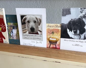 May include: A wooden card holder displaying several greeting cards. The cards feature winter and Christmas themes, including images of mittens, dogs, and religious scenes. Text includes "Warm Winter Wishes" and "Merry Christmas and Happy New Year!"
