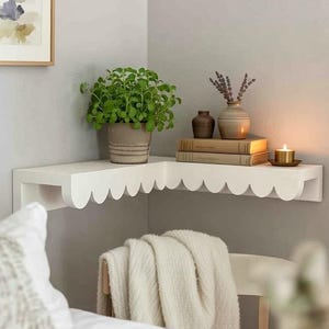 May include: A white L-shaped corner shelf featuring a decorative scalloped edge design. The shelf is mounted on a wall and holds a green potted plant, two stacked books, a small brown vase, a larger beige vase with dried lavender, and a lit candle.