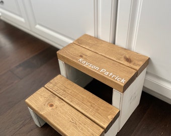 May include: A two-step wooden stool featuring a natural brown wood grain finish on the treads and white painted sides. The top step is personalized with the white text Kayson Patrick printed on the right side of the wooden surface.