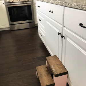 Wooden step stool, dark walnut stained steps with pink painted supports, personalized name hand painted on step, step stool in a kitchen, step stool against cabinet