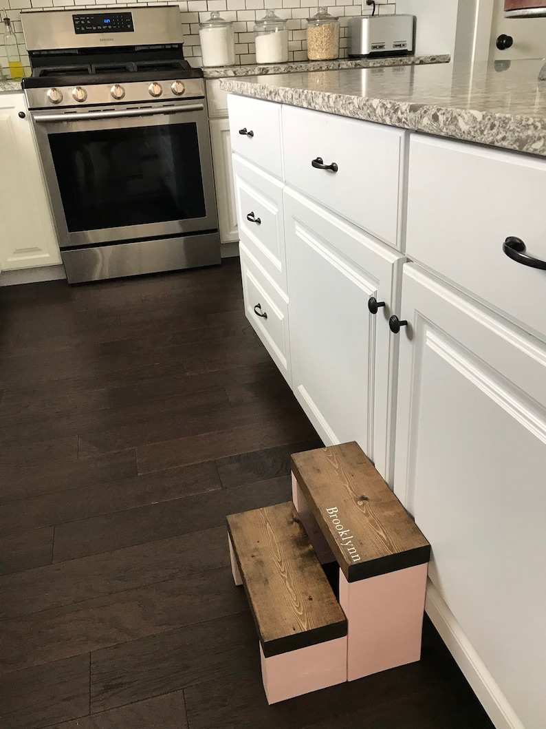 Wooden step stool, dark walnut stained steps with pink painted supports, personalized name hand painted on step, step stool in a kitchen, step stool against cabinet