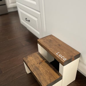 Wooden step stool, dark walnut stained steps with white painted supports, personalized name hand painted on step, step stool in a kitchen, step stool against cabinet