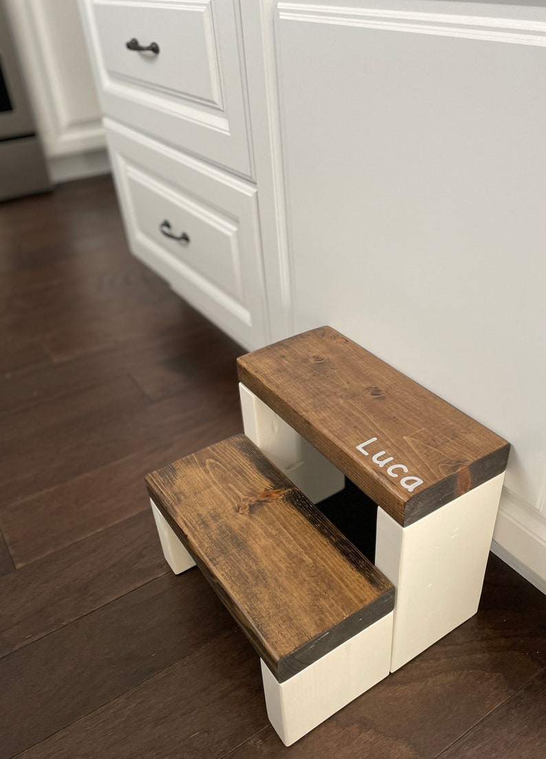 Wooden step stool, dark walnut stained steps with white painted supports, personalized name hand painted on step, step stool in a kitchen, step stool against cabinet