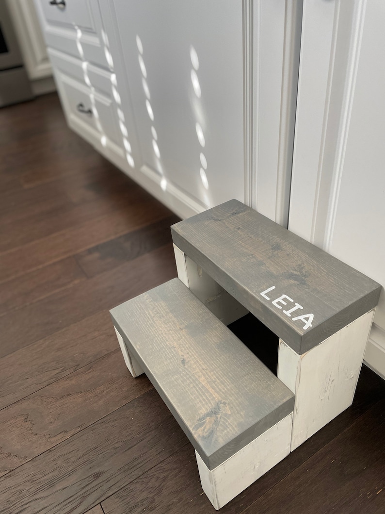 Wooden step stool, weathered gray stained steps with rustic white painted supports, personalized name hand painted on step, step stool in a kitchen, step stool against cabinet