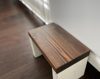 May include: A rectangular wooden step stool featuring a dark brown stained top surface with visible wood grain patterns and two solid white painted legs. The stool has a simple, functional design with a flat top and sturdy vertical supports.