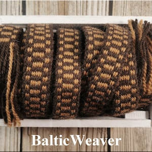 Op de afbeelding: Vijf geweven banden in bruin en beige met franjes aan de uiteinden in een witte houten doos. De doos heeft de tekst "Baltic Weaver" op de voorkant.