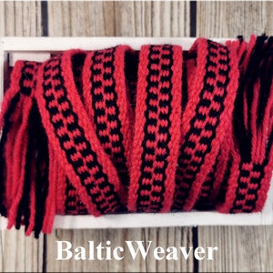 Op de afbeelding: Een close-up van verschillende rood-zwarte geweven banden met franjes aan de uiteinden. De banden zijn in een stapel gerangschikt en bevinden zich in een witte doos. De tekst "Baltic Weaver" is onder de doos gedrukt.