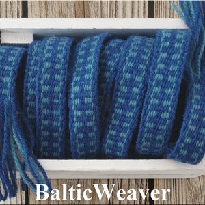 Op de afbeelding: Een close-up van blauw en turquoise geweven stof met franjes aan de uiteinden. De stof is opgerold in een witte houten doos. De tekst "Baltic Weaver" is onder de doos gedrukt.