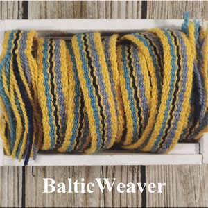 Op de afbeelding: Een close-up van een spoel geweven garen in een witte doos. Het garen is een mix van geel, blauw, grijs en zwart. De tekst "Baltic Weaver" is onder de doos gedrukt.