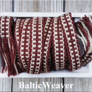 Op de afbeelding: Een close-up van een geweven rood en wit lint met franjes aan de uiteinden. Het lint zit in een witte houten doos. De tekst "Baltic Weaver" is onder de doos gedrukt.